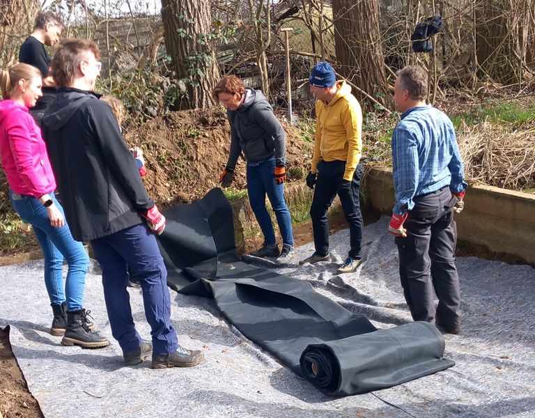 Die Bild zeigt eine Gruppe von Menschen bei Bau- oder Wartungsarbeiten. Die Szene ist in einer natürlichen Umgebung mit Bäumen im Hintergrund. Die Personen tragen Freizeitkleidung, darunter Jacken, Hüte und Handschuhe, da das Wetter kühl oder nass ist. In der Vordergrund befindet sich ein großes schwarzes Tarp oder Abdeckung auf dem Boden, das eine flache, rechteckige Materialstück ist. Das Tarp ist gerollt und auf einer Sandfläche platziert, was darauf hindeutet, dass es für die Abdeckung oder Schutz eines Untergrundes verwendet werden könnte. Die Gruppe besteht aus mindestens fünf Personen, wobei eine Person im Zentrum eine gelbe Jacke trägt und eine andere Person auf der rechten Seite ein blaues Plaid-Shirt trägt. Die Person in der gelben Jacke scheint das Tarp zu handhaben, während die anderen beobachten oder helfen. Die Person auf der linken Seite trägt eine schwarze Jacke und scheint das Tarp zu betrachten, möglicherweise über oder eine Planung für die Aktivität zu diskutieren.